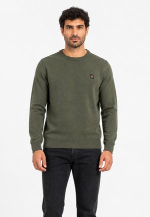 Uomo con capelli corti e ricci che indossa un maglione verde oliva con scollo tondo e jeans neri, in piedi davanti a uno sfondo bianco semplice.