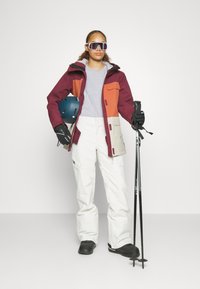 Lyžiarsky outfit: bordový a oranžový sako s sivými akcentmi, biele nohavice, čierne rukavice, lyžiarske palice a modrá helma. Veľké slnečné okuliare a športový dizajn.