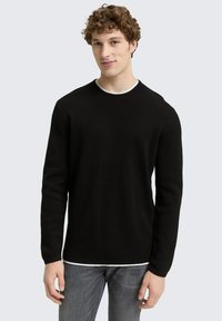 Schwarzer, gerippter Pullover mit langen Ärmeln und rundem Halsausschnitt, mit kontrastierendem weißen Besatz. Über grauen Jeans getragen.