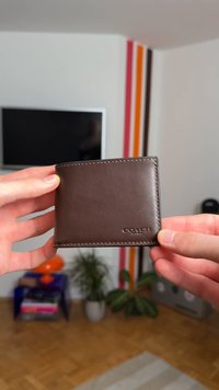 Roke v rokah držita rjavo usnjeno denarnico znamke Coach v dnevni sobi s televizorjem, barvnimi stenskimi črtami, rastlinami in knjigami v ozadju.