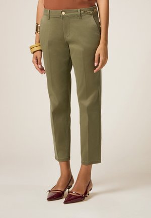 Olijfgroene chino's met een rechte pasvorm, voorzien van een voorzak en een decoratieve kettingaccent op de tailleband. Draagt met kastanjebruine hakken.