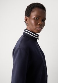 Navy zip-up jacket with a high collar and white striped accents. Smooth texture with a fitted silhouette, featuring a silver zipper detail.