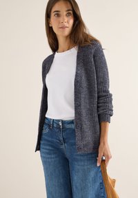 Laivastonsininen neulecardigan, jossa on ribbaustekstuuri ja viisi nappia edessä. Päällä valkoinen paita ja siniset farkut.