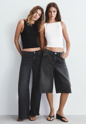 Két nő áll egy fehér fal előtt, ujjatlan felsőket és laza fekete farmer alsókat viselve, szandálokkal.