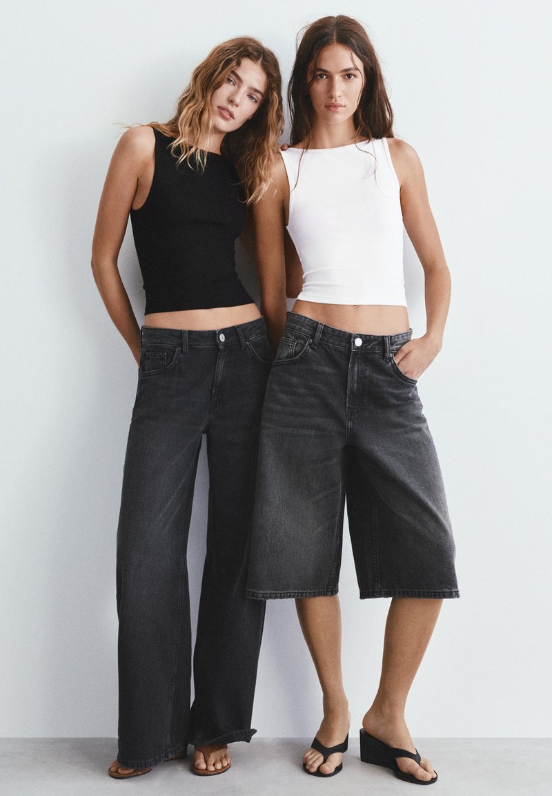 Deux femmes se tiennent contre un mur blanc, portant des hauts sans manches et des pantalons en denim noir larges avec des sandales.