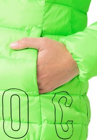 Chaqueta acolchada de color verde neón con una mano en el bolsillo lateral. Presenta una textura acolchada y letras impresas en negro en la parte frontal.
