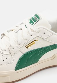 Fehér bőr sportcipő zöld díszítéssel, arany "Puma" logóval, lapos fehér fűzőkkel és mintázott gumitalppal.