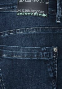 Mörkblå denimjeans med en slät textur, som har en bakficka med en silverknapp och en svart etikett som säger "clear focus."