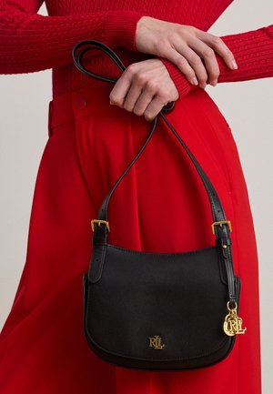 Personne portant un pull rouge et un pantalon tient un sac à main en cuir noir avec un logo RL doré et un charm par ses anses derrière son dos.