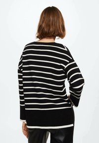 Vrouw met kort bruin haar, gekleed in een zwart-witte horizontaal gestreepte trui en zwarte broek, staat met één hand op haar heup.