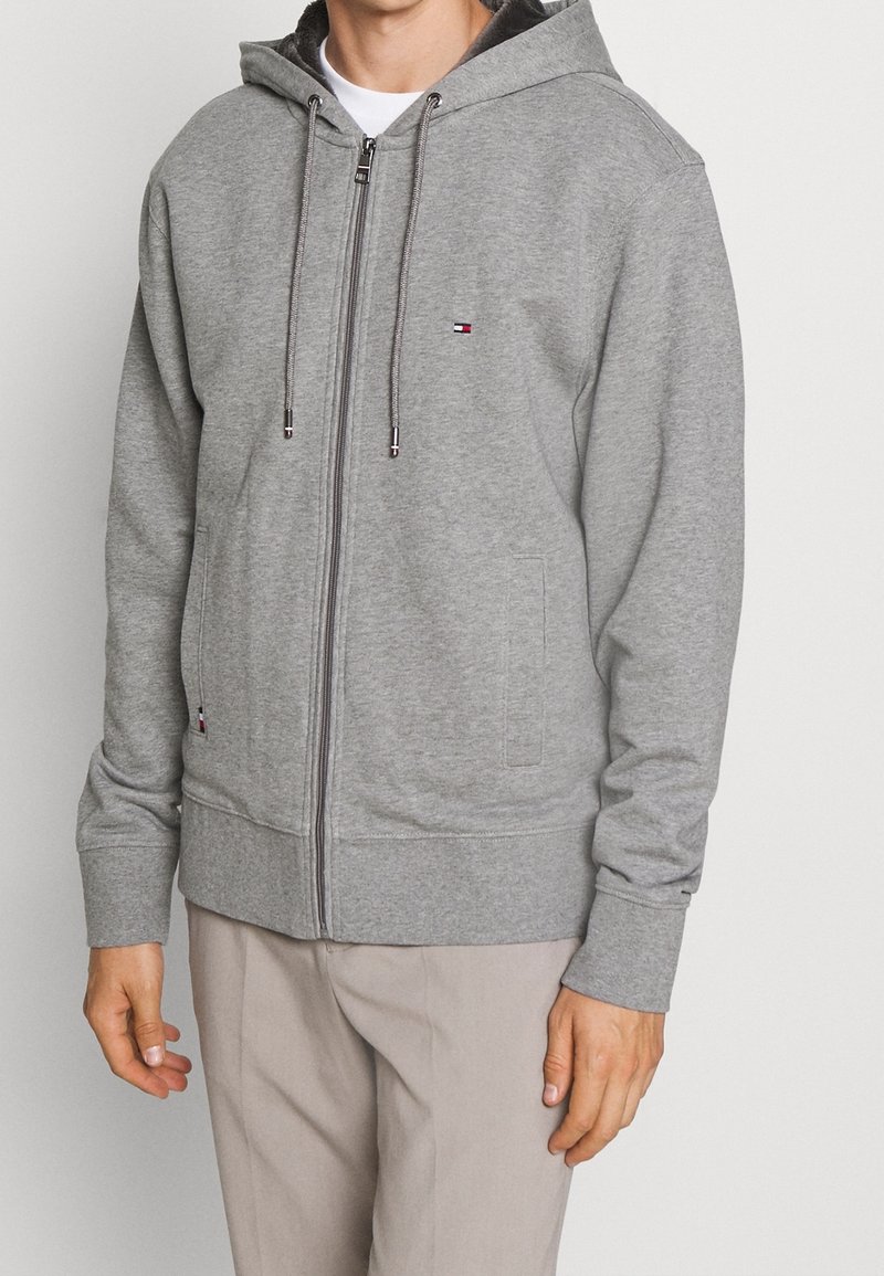 Homme portant un sweat à capuche gris clair zippé avec cordons et un pantalon beige, debout devant un fond uni clair.