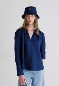 Camicia in lino blu navy a maniche lunghe con scollo a V, aperture laterali e tessuto leggermente strutturato, abbinata a un cappello a secchiello blu navy con logo.