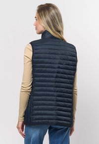 Oblečena je podložena brezrokavnik temno modre barve z visokim ovratnikom, prešitim vzorcem in gladko teksturo. Oblečena je čez bež dolg rokav.