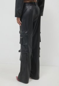 Rückansicht einer Person, die schwarze, weite Cargo-Hosen aus Kunstleder mit mehreren Klappentaschen und roten spitzen Schuhen trägt.