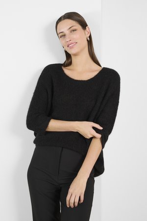 Schwarzer Strickpullover mit lockerer Passform, weitem Ausschnitt und Dreiviertelärmeln, kombiniert mit hochtaillierten schwarzen Hosen. Minimalistisches Design.