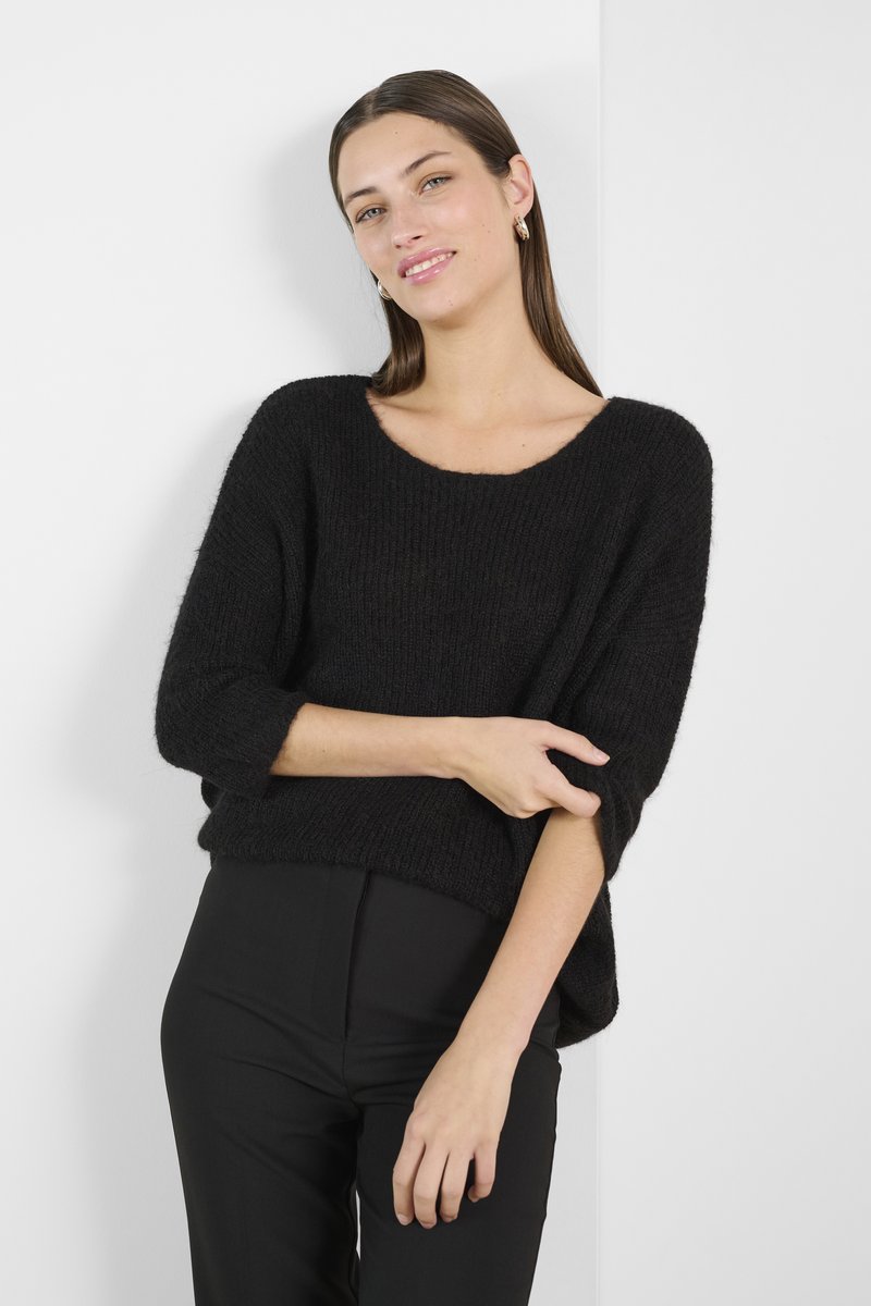 Melnā adīta džemperis ar brīvu piegriezumu, plašu kakla izgriezumu un trīs ceturtdaļu piedurknēm, kombinēts ar augsto vidukļa melno biksēm. Minimālisma dizains.