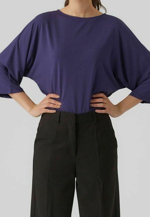 Personne portant un haut bleu foncé ample à larges manches et un pantalon noir taille haute, les mains posées sur les hanches.