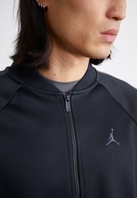 Sort zip-up jakke lavet af glat stof. Den har en ribbet krave og et lille gråt Jordan-logo på brystet over lynlåsen.