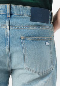 Světle modré džíny z denimu s oranžovým prošitím, které mají koženou značku Lacoste a malé vyšívané logo na zadní kapse.