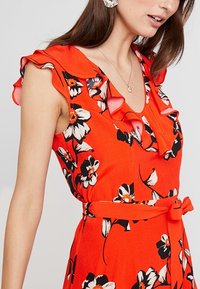 Robe à fleurs rouge avec des fleurs noires et blanches, des manches à volants et une ceinture assortie à la taille. Tissu doux avec une texture légère.