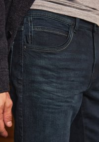 Tmavě modré džínové kalhoty s tapered střihem, s žlutým prošitím podél kapes a švů, a s mírnou texturou viditelnou ve tkanině.