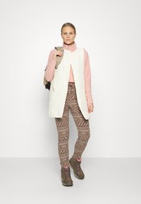 Colete acolchoado branco por cima de uma blusa de mangas longas rosa, combinado com leggings castanhas com padrão e botas de montanha castanhas. A segurar uma mochila bege.