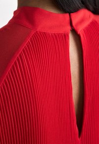 Rode geplooide blouse met een hoge halslijn en een keyhole achterkant, gemaakt van een gladde stof met gestructureerde verticale lijnen.