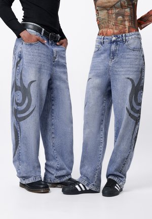 Baggy blue denim jeans with black tribal designs on the sides, featuring visible stitching and belt loops. Worn with contrasting footwear.