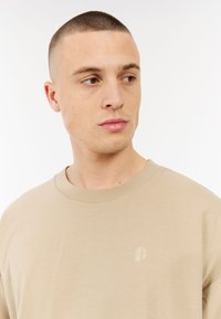 Beige katoenen t-shirt met een ronde halslijn en korte mouwen. Bevat een geborduurd logo op de borst. Glad materiaal, ontspannen pasvorm.