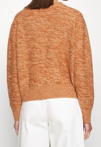 Osoba v měkkém oranžovém pleteném svetru a bílých kalhotách, zobrazená zezadu na jednobarevném pozadí.