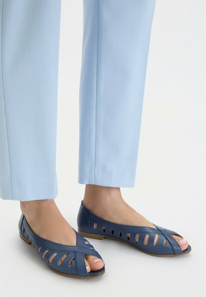 Füße in hellblauen, verkürzten Hosen und dunkelblauen, offenen flachen Schuhen mit Cut-out-Details vor weißem Hintergrund.