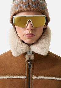 Skibrille mit einem weißen Rahmen und gelben Gläsern, getragen zu einer braunen Jacke mit Schafslederen Kragen, die einen Reißverschluss mit einem Logo-Detail aufweist.
