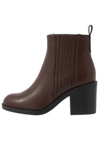 Brązowy botek na kostkę z faux skóry, z okrągłym noskiem, żebrowanym detalem po bokach i masywnym czarnym obcasem. Pętla z tyłu ułatwiająca zakładanie.