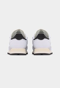 Ζευγάρι λευκών χαμηλών αθλητικών παπουτσιών με μαύρες ταμπέλες στη φτέρνα και μπεζ ενδιάμεσα σόλες, σε θέα από πίσω σε λευκό φόντο.