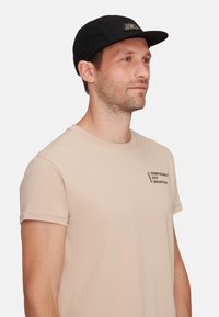 Svart keps med platt skärm, prydd med en logopatch. Bäraren har på sig en ljusbeige t-shirt med detalj av tryckt text på bröstet.
