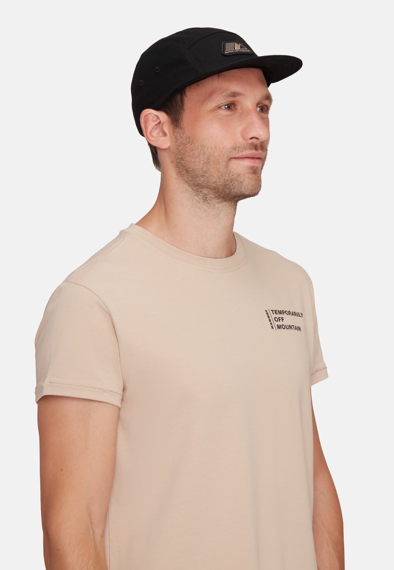 Svart keps med platt skärm, prydd med en logopatch. Bäraren har på sig en ljusbeige t-shirt med detalj av tryckt text på bröstet.