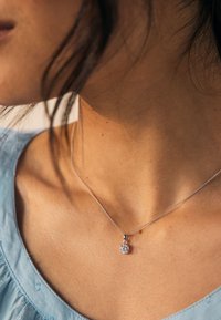 Silberne Halskette mit einer floralen Anhänger, versehen mit einem klaren, funkelnden Edelstein, getragen vor einem hellblauen Stoffhintergrund.