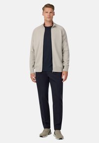 Jasnoszara bluza zapinana na zamek, noszona na granatowej koszulce, w połączeniu z ciemnymi spodniami i jasnoszarymi sneakersami. Prosty design, gładka tekstura i brak widocznych wzorów.