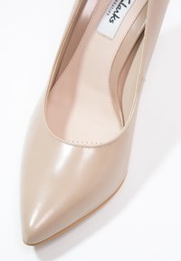 Chaussure à talons aiguilles en cuir beige avec bout pointu, avec des coutures visibles et une semelle brun clair sur un fond blanc.