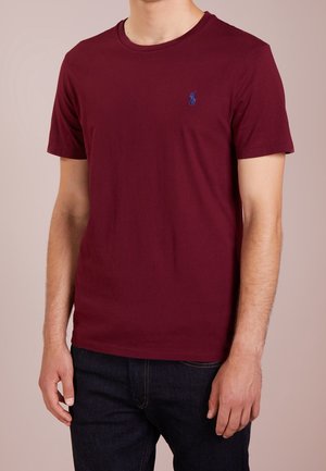 Burgundowa bawełniana koszulka z okrągłym dekoltem i krótkimi rękawami; posiada małe niebieskie logo na piersi; gładka tekstura i klasyczny krój.