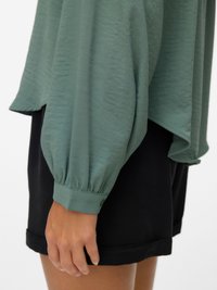Green textured blouse with dropped shoulders and long puff sleeves, paired with black shorts. Blouse has a rounded hem and buttoned cuffs.