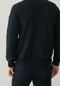 Uomo che indossa un felpa nera a maniche lunghe e pantaloni cargo neri, in piedi con le mani rilassate ai lati, di spalle rispetto alla fotocamera.
