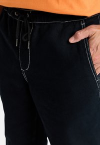 Črne hlače z pasom na vrvico, ki imajo bele šive in stranske žepke. Tkanina se zdi vzdržljiva z teksturirano površino.