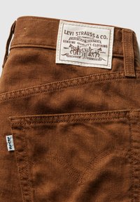 Hnedé džínsy z bavlnenej tkaniny s výrazným prehnutým švom, značka Levi Strauss & Co. na páse a malá biela etiketa.