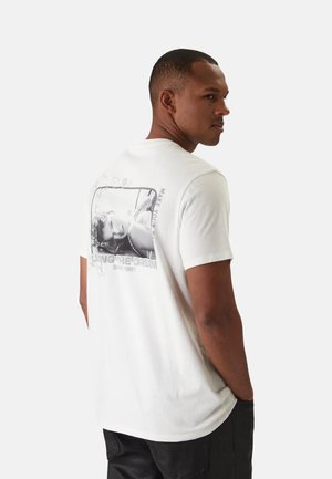 Uomo con maglietta bianca con foto in bianco e nero e testo "Living the Dream Since 1981" sul retro, che guarda oltre la spalla.