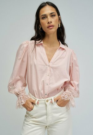 Femme portant une chemise légère rose pâle à boutons avec des poignets en dentelle et un pantalon blanc taille haute, debout les mains dans les poches devant un fond uni.
