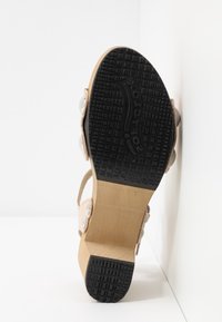 Sandale avec talon plateforme en bois et semelle en caoutchouc noir, montrant un motif de grip texturé et le nom de la marque en dessous.