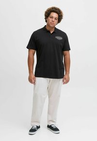 Schwarzes Poloshirt mit einem Logo auf der Brust, kurzen Ärmeln und einem klassischen Kragen, kombiniert mit hellbeigen Hosen und schwarzen Sneakers.