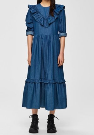 Jonge vrouw die een lange blauwe denimjurk draagt met ruches aan de kraag en zoom, opgerolde mouwen en zwarte veterlaarzen, staand tegen een witte achtergrond.