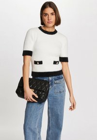 Suéter blanco de manga corta con ribetes negros y bolsillos, presenta una textura esponjosa. Jeans de tiro alto casuales y un bolso negro acolchado.